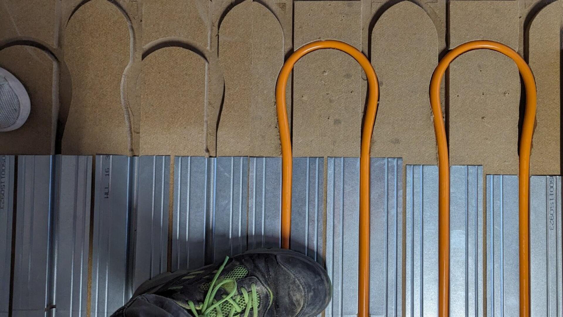 A person wearing a black shoe with green laces stands on metal panels next to orange heating pipes arranged in loops and cutouts in a brown surface above.