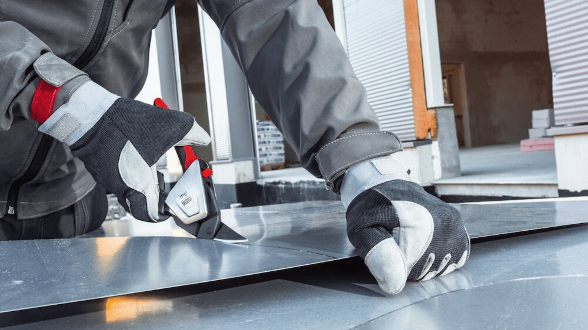 Eine Person mit Schutzhandschuhen und Jacke schneidet mit einem Universalmesser im Freien ein Metallblech durch, in der Nähe eines Gebäudes mit offenen Garagentoren im Hintergrund.
