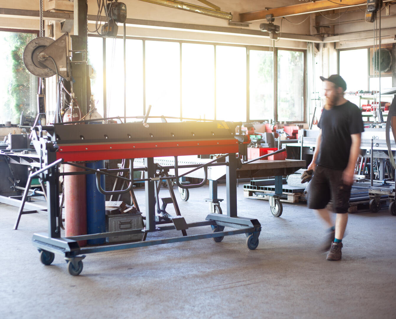 A man in a cap and dark clothes walks through a bright workshop full of equipment, including a large metal machine on wheels in the center and various tools on benches by the windows.
