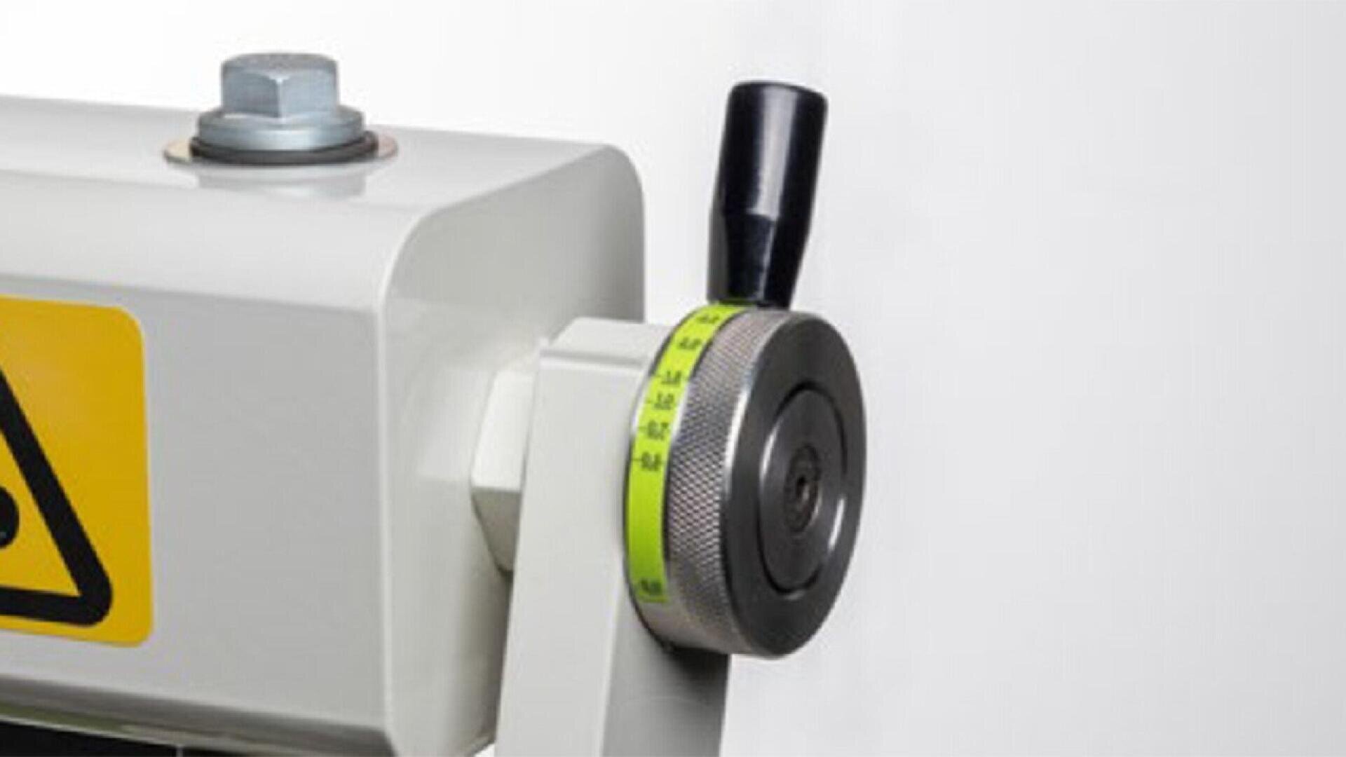 Close-up of a metal machine adjustment knob with a green and gray numbered dial, a black handle, and a partially visible yellow warning sign on a white background.