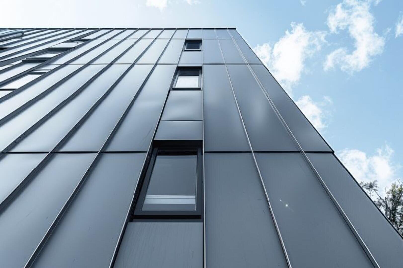 View from a low height of a modern building with smooth metal panels and several narrow windows against a blue sky with scattered clouds.