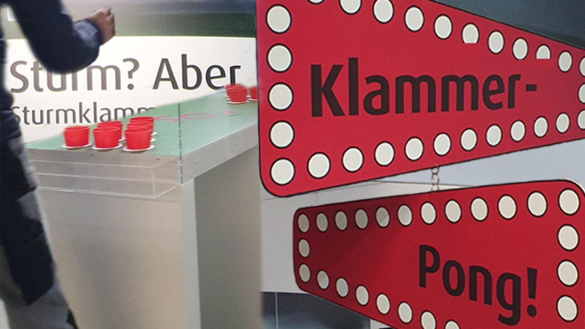 A person plays a game with red plastic cups on a table. In the foreground, there are red dotted signs reading Klammer-Pong! and a background sign with partial German text Sturm? Aber.