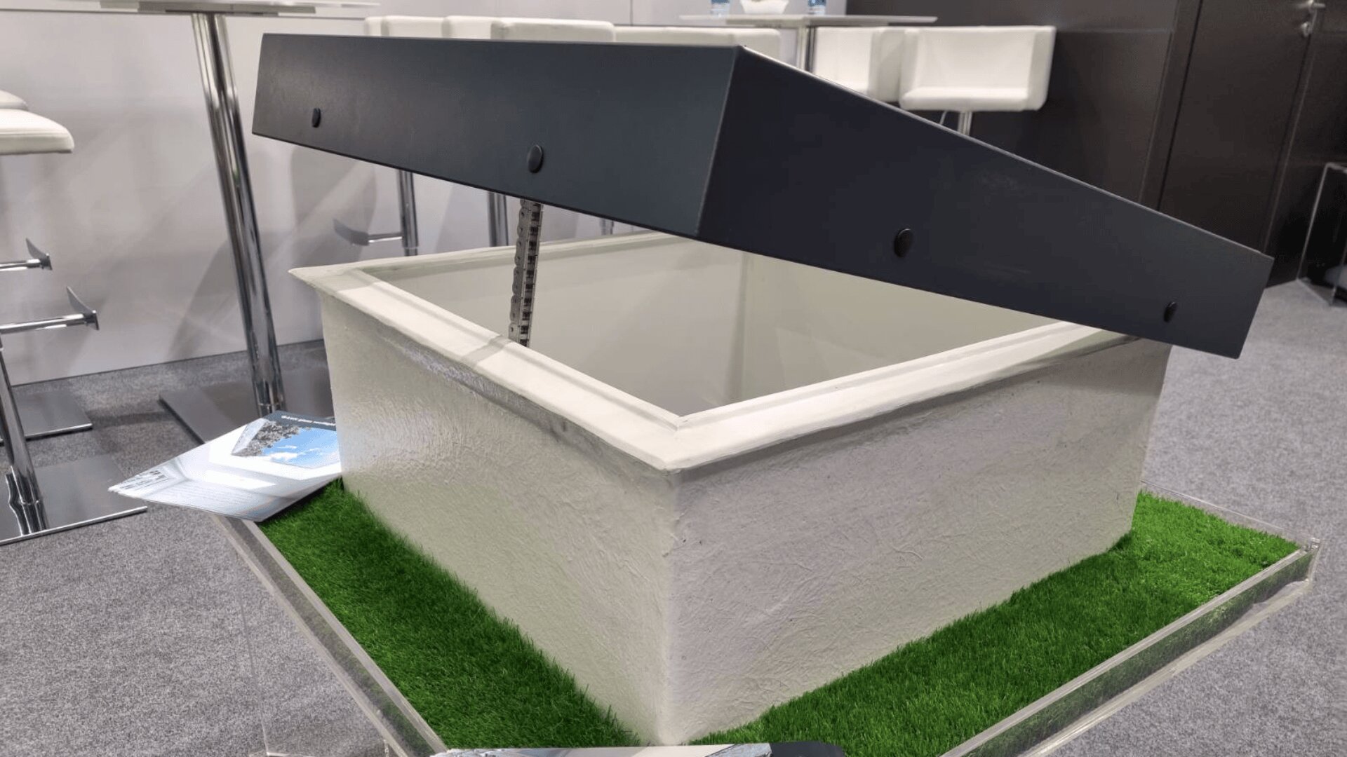 A white rectangular box with a hinged black lid is displayed on a base of artificial grass. The box is partially open, revealing the interior, and stands on a platform in an indoor setting with modern white chairs in the background.
