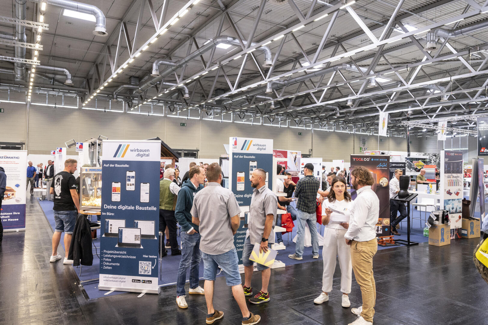 Auf einer belebten Indoor-Messe oder -Ausstellung tauschen sich die Menschen aus und stöbern in den Ständen. Verschiedene Auslagen, Banner und Informationsstände sind unter einer großen industriellen Decke mit hellen Lichtern zu sehen.