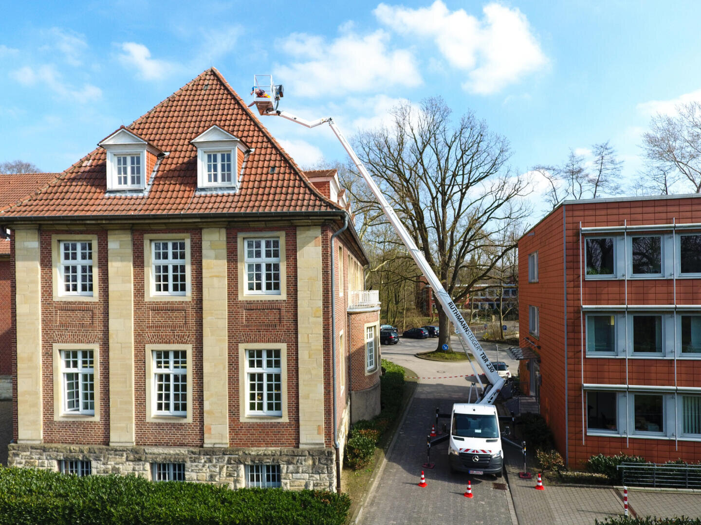 Eine Person steigt mit einem Hubsteiger auf das Dach eines Backsteingebäudes mit rotem Ziegeldach und arbeitet in der Nähe von Dachfenstern. Ein Lkw mit ausgefahrener Hebebühne ist darunter geparkt, und orangefarbene Kegel versperren den Bereich.