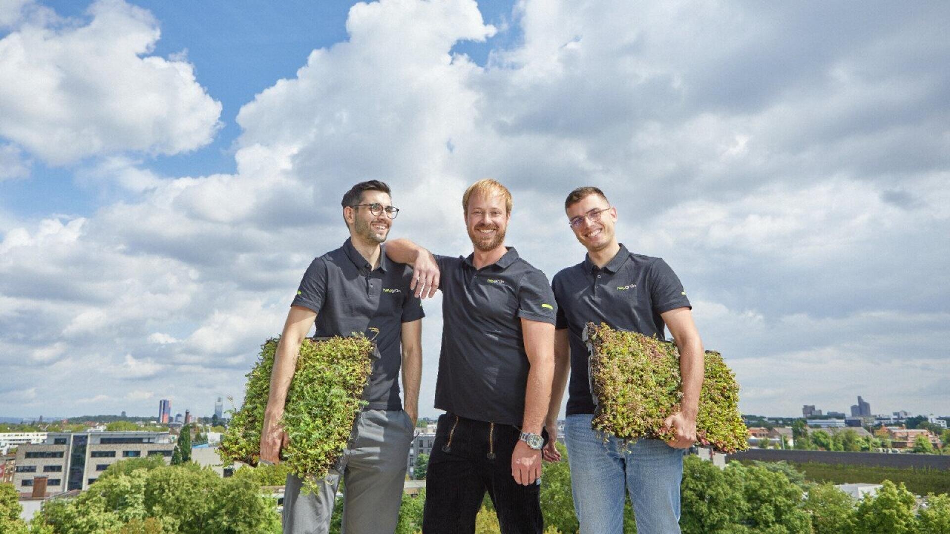 Three men in black shirts stand on a rooftop, smiling and holding rectangular frames filled with green plants. A cityscape and cloudy sky are visible in the background.