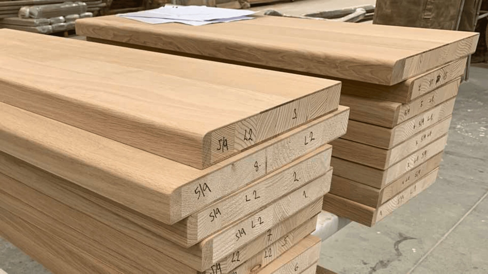 Ein Stapel fein säuberlich zugeschnittener Holzbretter mit Zahlen und Buchstaben an den Enden, die auf einer Werkbank in einer Werkstatt liegen. Oben auf den Brettern liegen ein paar Blätter Papier.