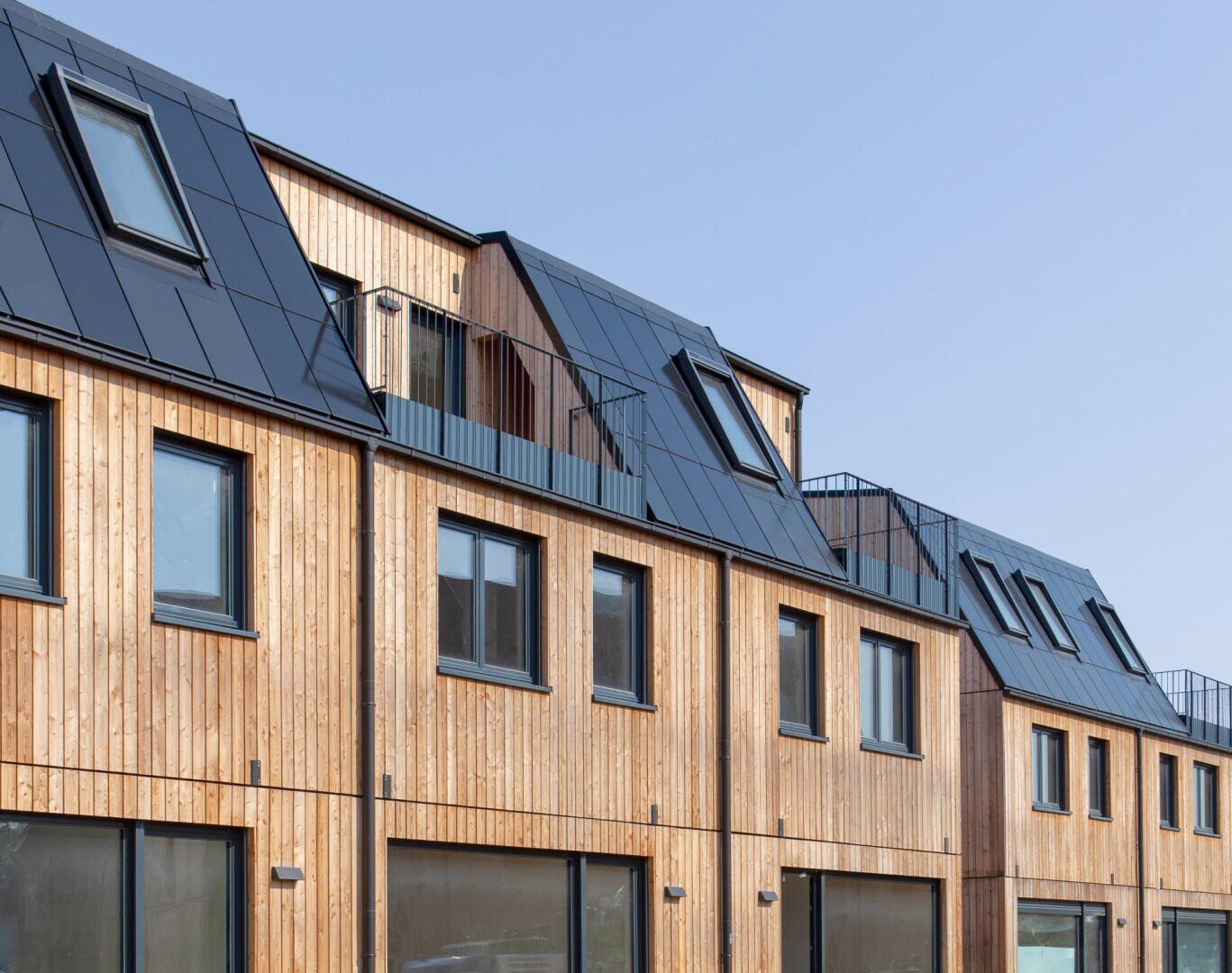 A row of modern townhouses with wooden facades and black metal roofs with lots of windows and skylights against a clear blue sky.