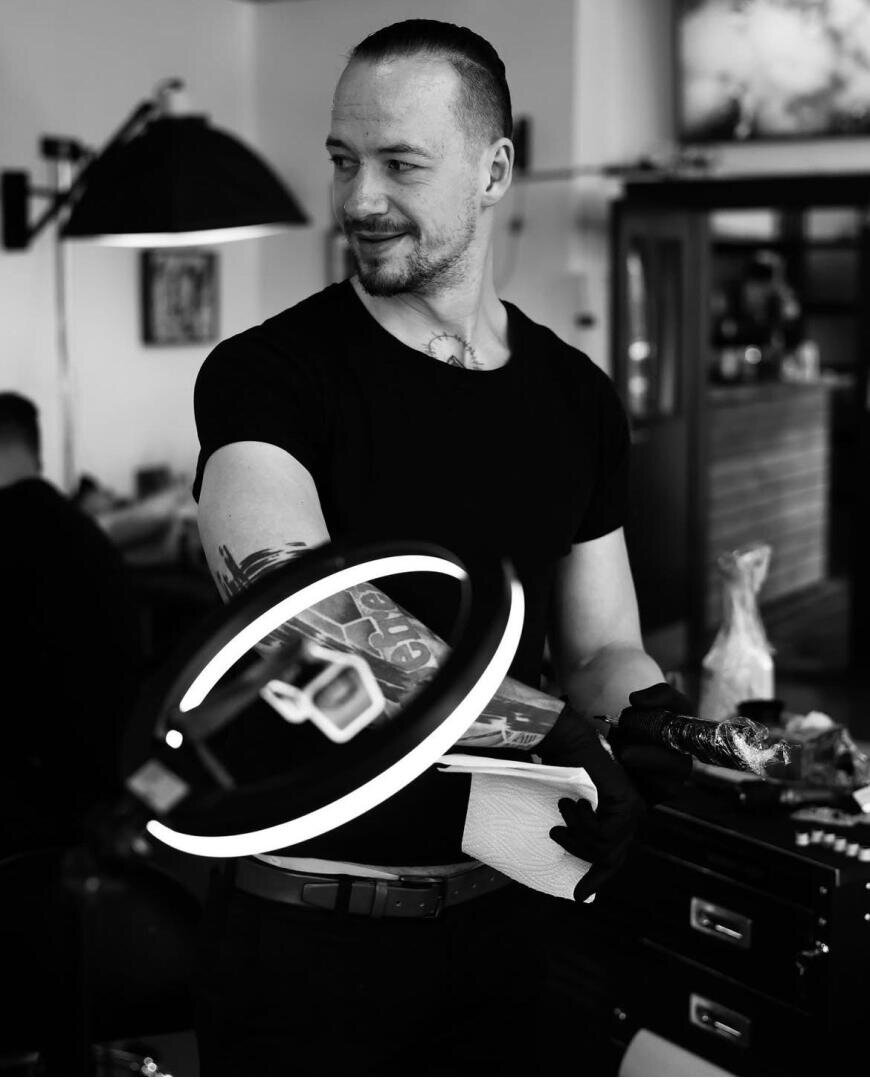 Ein Tätowierer mit einem tätowierten Arm steht in einem Studio, hält ein Papiertuch in der Hand und trägt Handschuhe. Ein Ringlicht beleuchtet seinen Arm, und er blickt lächelnd zur Seite. Das Bild ist in schwarz-weiß gehalten.