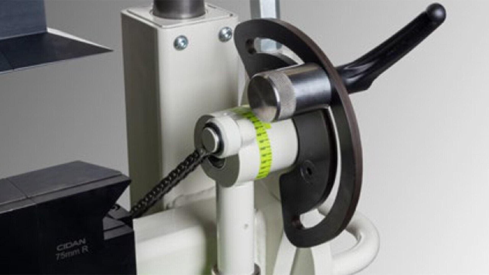 Close-up of a mechanical device featuring a handle, a cylindrical metal component, a circular scale with green markings, and a chain. The device appears to be part of industrial or laboratory equipment.