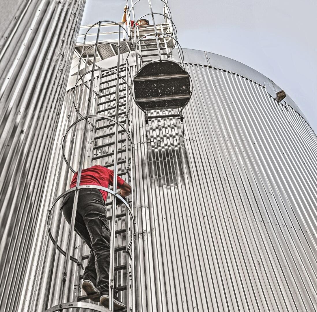 A person wearing a red shirt and dark pants climbs a caged metal ladder attached to the side of a large, silver, corrugated industrial silo, next to which another ladder and another silo are visible.
