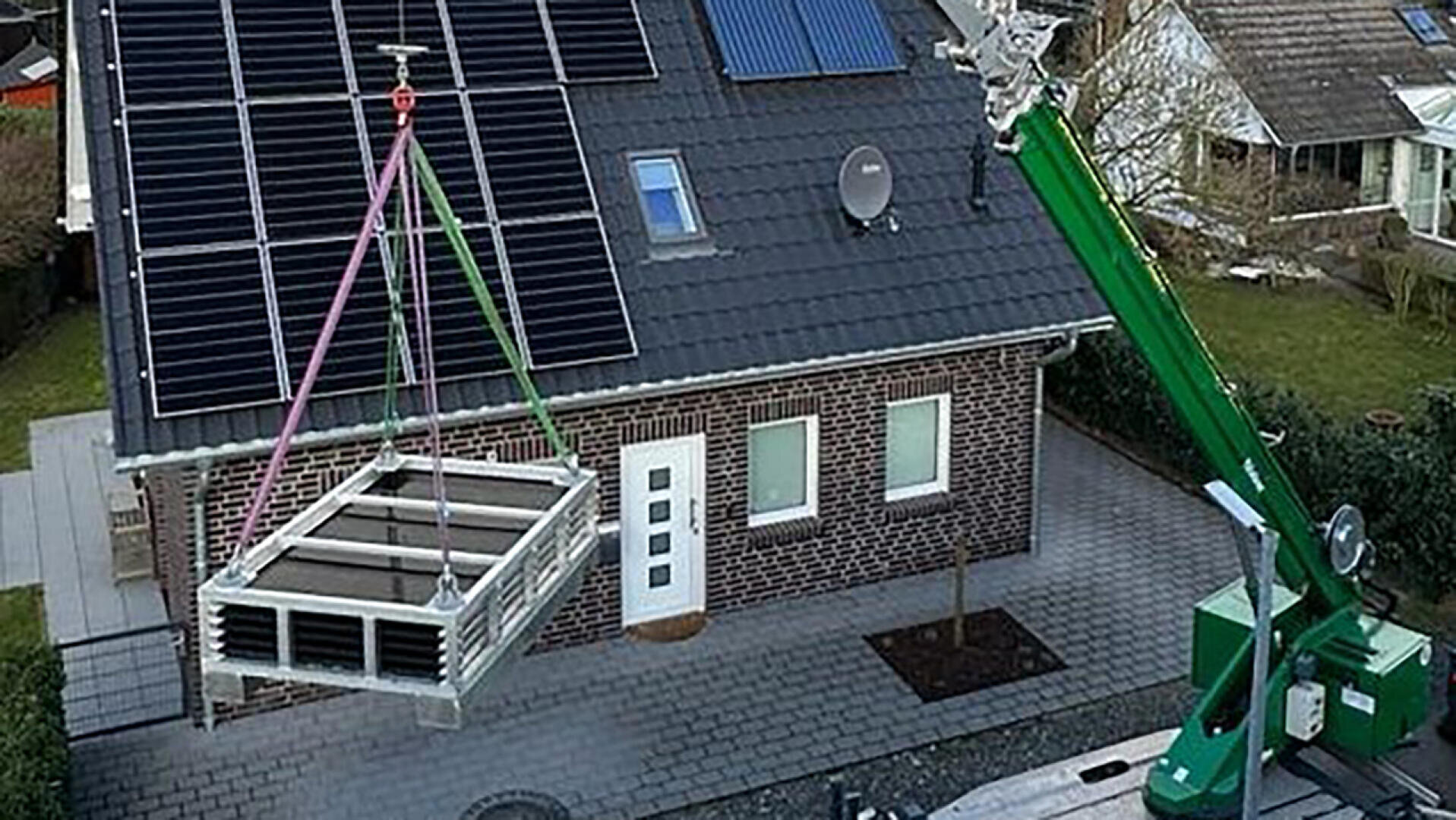 Ein Kran hebt einen modularen Abschnitt auf das Dach eines Backsteinhauses mit Solarmodulen, weiß gerahmten Fenstern und einem gemusterten Gehweg davor. Die Szenerie wirkt wohnlich und gepflegt.