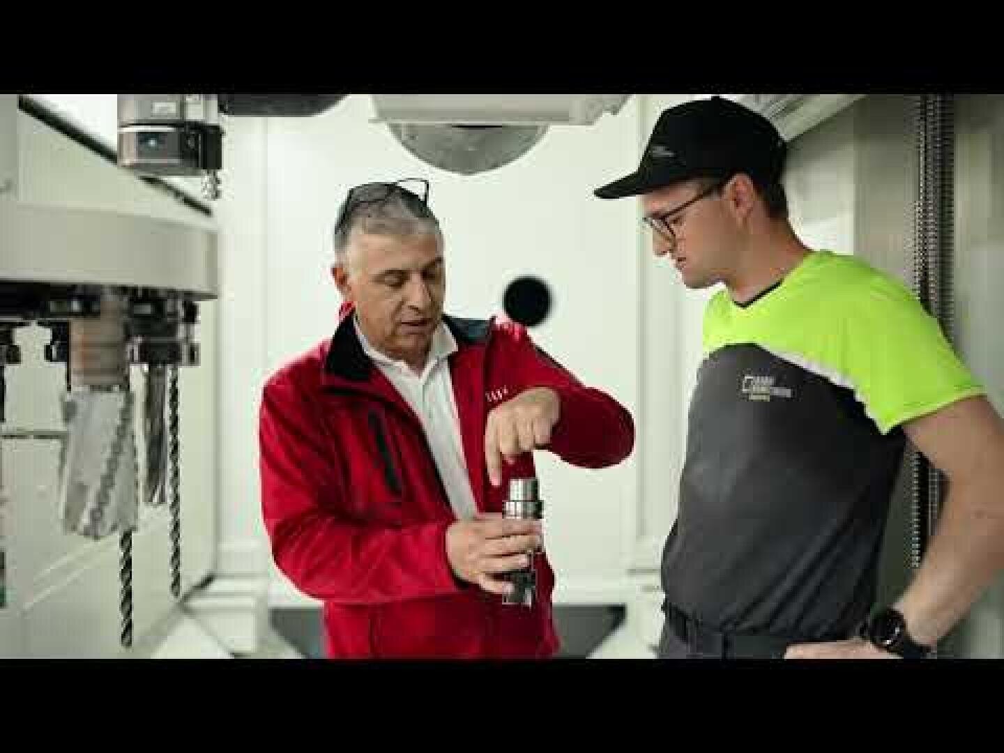 Zwei Männer stehen in einer Werkstatt; der eine in einer roten Jacke erklärt dem anderen, der eine Kappe und eine Schutzbrille trägt, ein Metallwerkzeugteil. Im Hintergrund sind Maschinen und Geräte zu sehen.