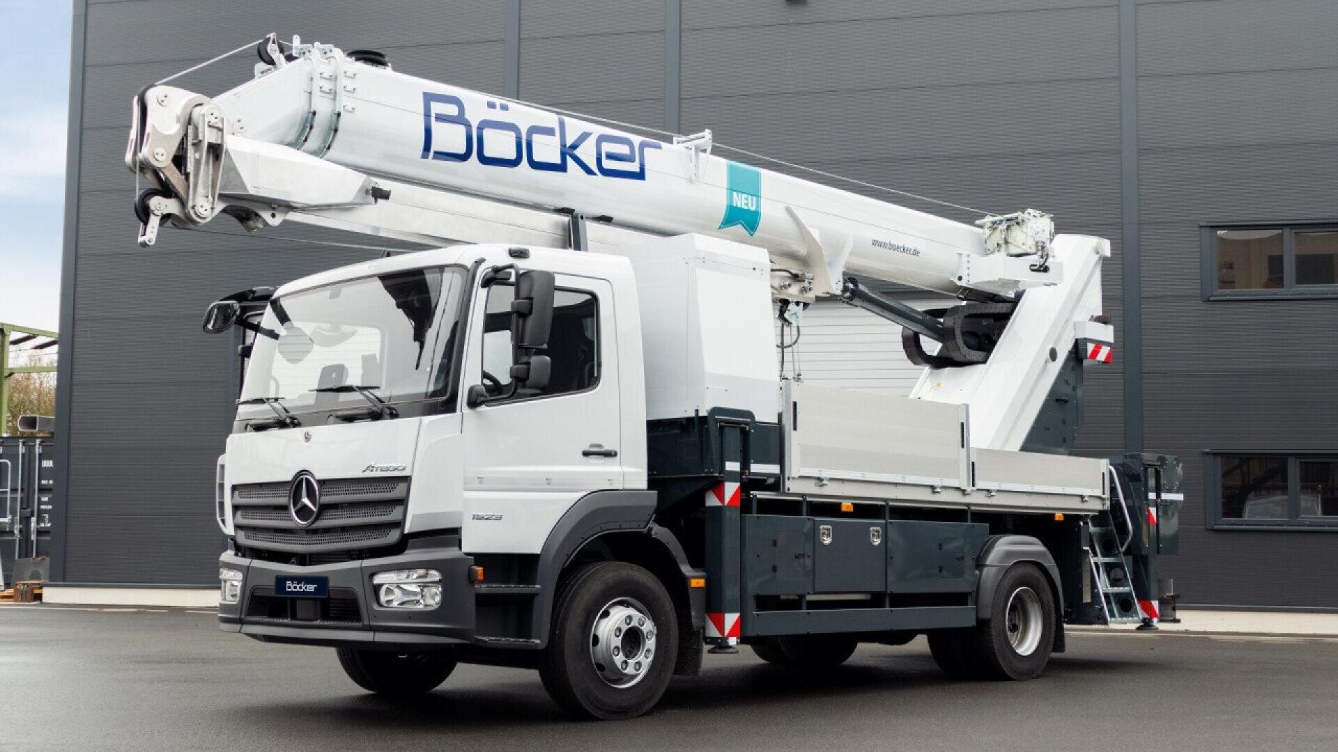 A white Böcker truck-mounted crane with an extended telescopic boom is parked on a paved surface in front of a gray industrial building.