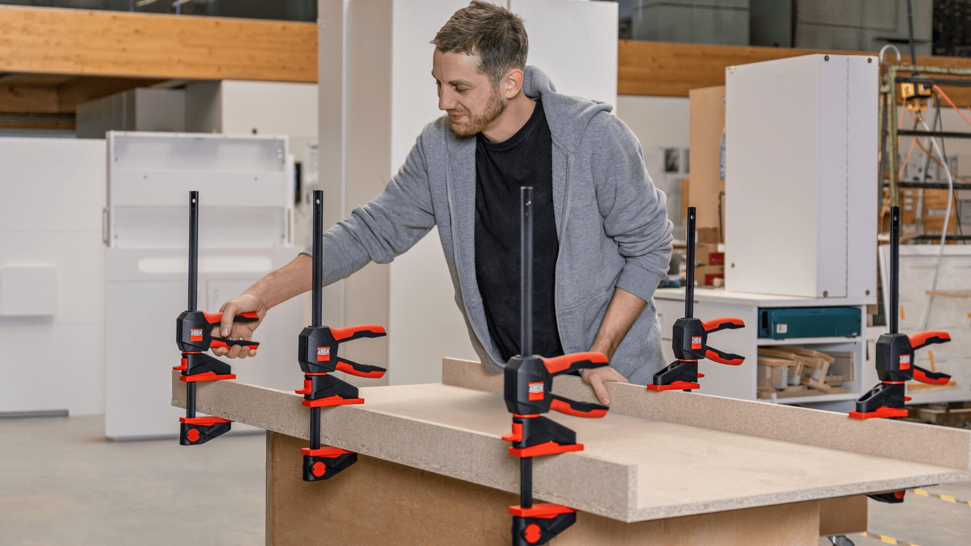 Eine Person in einer Werkstatt verwendet mehrere rote und schwarze Zwingen, um Holzstücke auf einer Werkbank zu befestigen, um ein Holzbearbeitungsprojekt vorzubereiten.