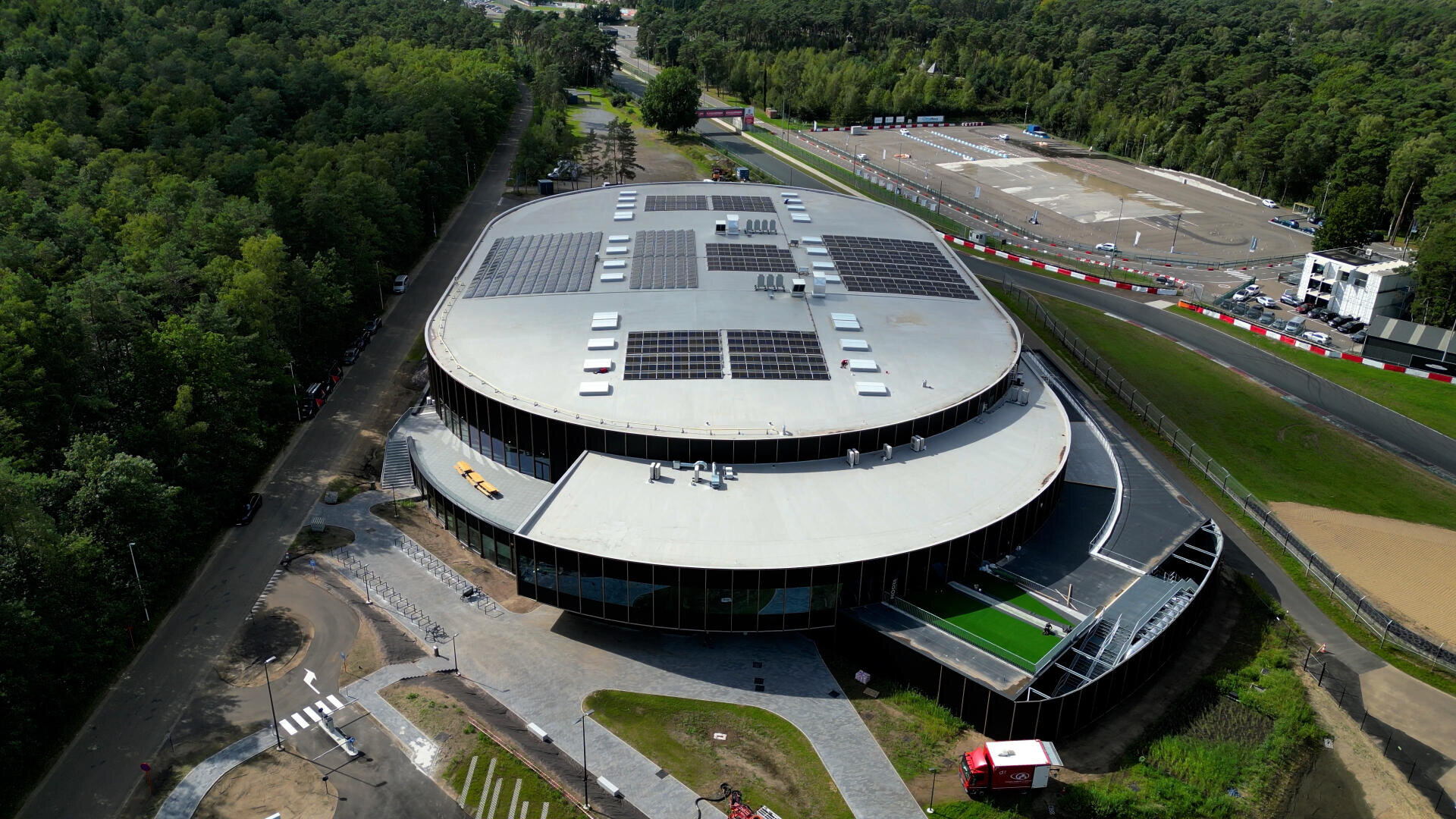 Aerial view of a large, modern round building with solar panels on the roof, surrounded by trees, roads and a nearby race track with red and white barriers.