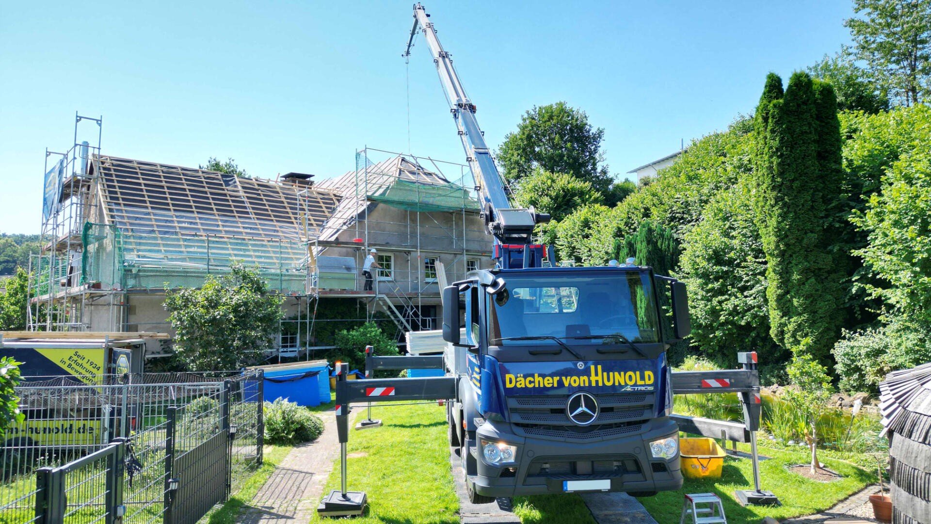 Ein großer Autokran mit der Aufschrift „Dächer von Hunold“ steht vor einem Haus mit Dachkonstruktion. Gerüst und Dachziegel sind sichtbar; üppiges Grün umgibt das Gelände.