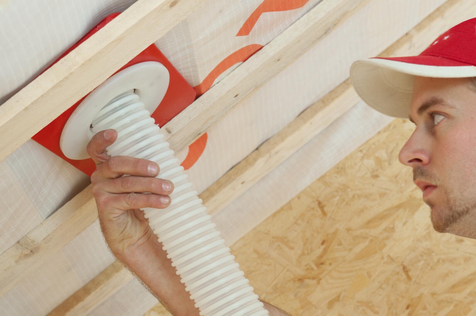 Ein Mann mit einer rot-weißen Kappe installiert ein flexibles weißes Lüftungsrohr durch eine Deckenkonstruktion aus Holzbalken und Holzspanplatten. Mit einer Hand befestigt er das Rohr in einer roten Montageplatte.