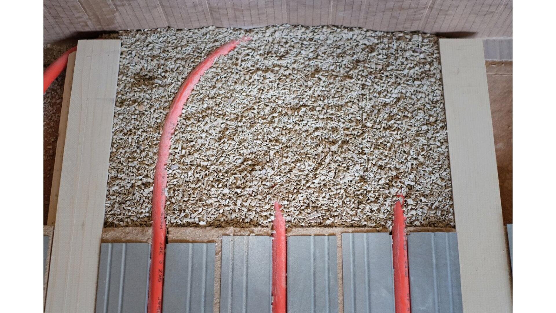 A close-up of a floor section showing pink underfloor heating pipes laid within a layer of insulation material and wooden framing. Metal plates are also visible, used for heat distribution.