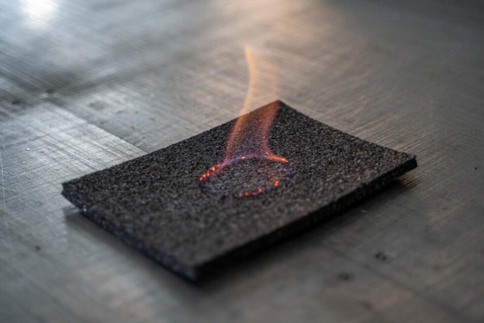 In der Mitte brennt ein rechteckiges Stück dunkles, strukturiertes Material mit einer kleinen orangefarbenen Flamme und Glut, die auf einer metallischen Oberfläche ruht.