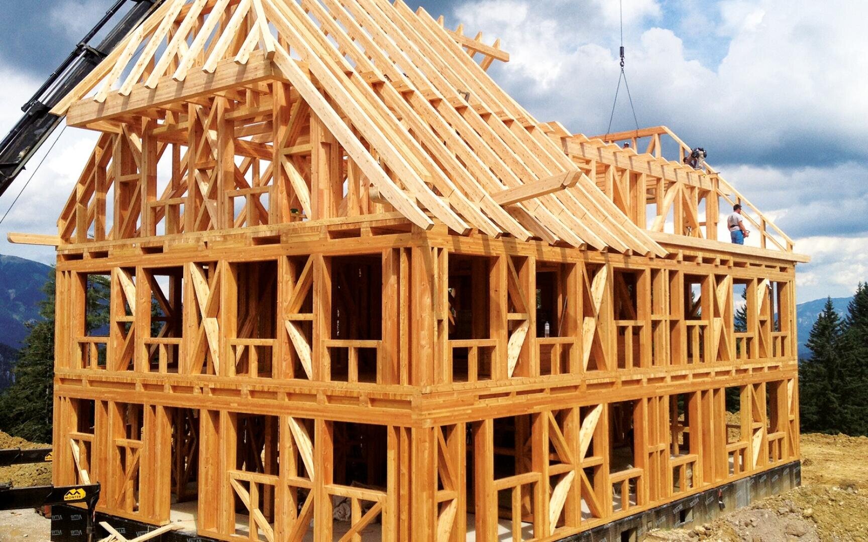 Ein im Bau befindliches zweistöckiges Holzhaus mit freiliegendem Holzskelett und Dachkonstruktion, mit Arbeitern im oberen Stockwerk und einem Kran zur Unterstützung. Das Gebäude befindet sich vor einem Hintergrund aus Bäumen und bewölktem Himmel.