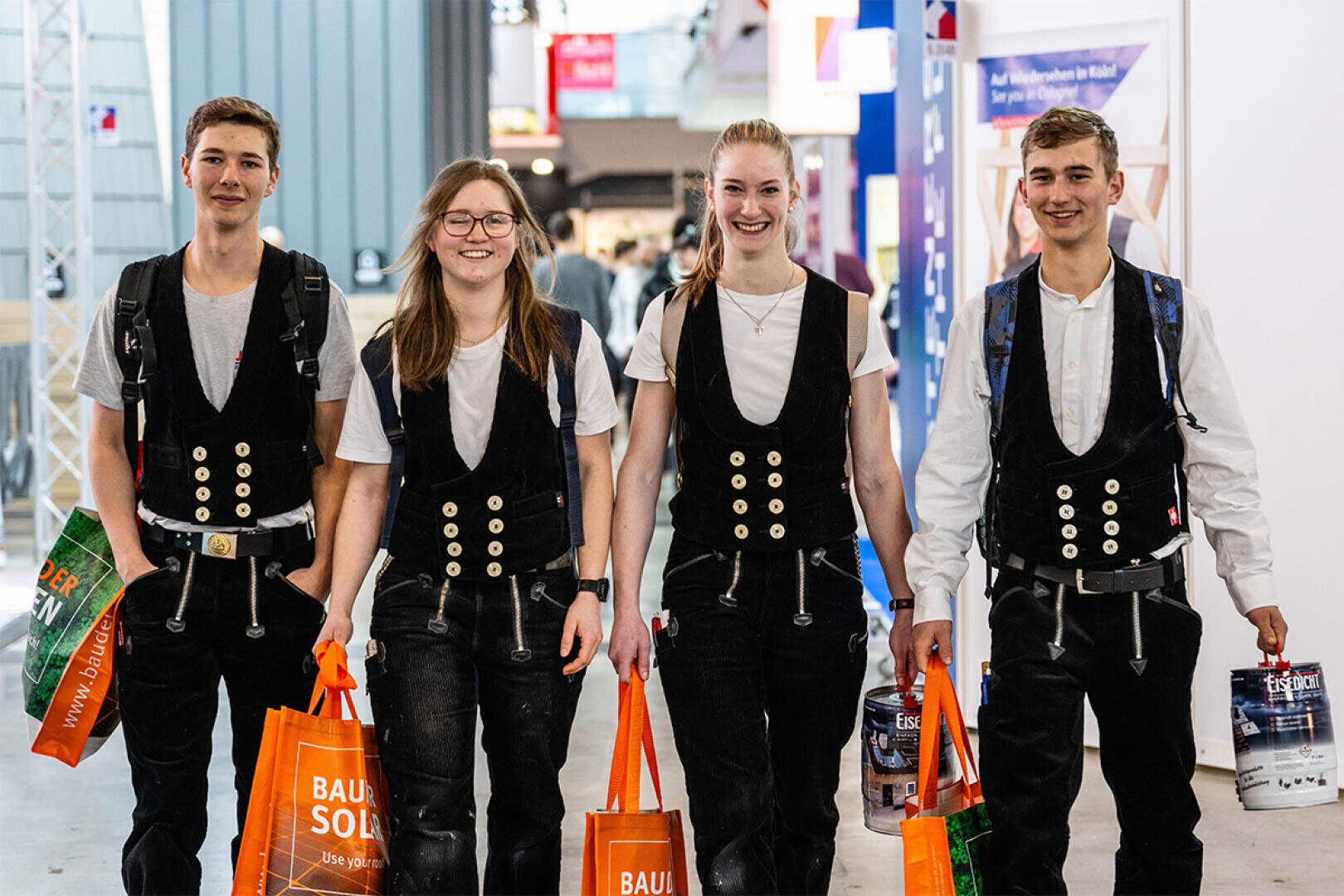 Vier junge Leute, die passende traditionelle schwarze Westen, weiße Hemden und dunkle Hosen tragen, gehen lächelnd und mit leuchtend orangefarbenen Tragetaschen in den Innenraum. Sie scheinen auf einer Veranstaltung oder Ausstellung zu sein.