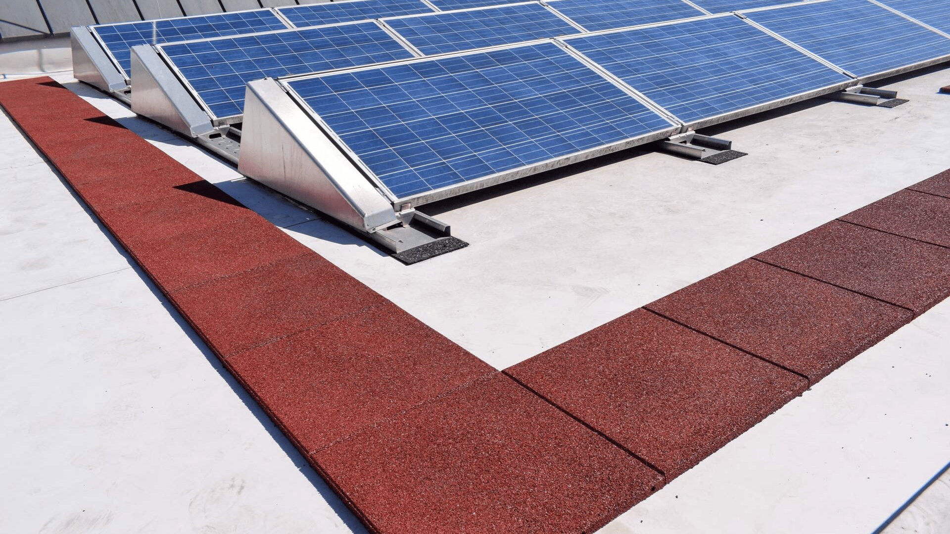 Auf einem flachen weißen Dach sind Reihen von blauen Solarmodulen angebracht, die neben einem Weg aus roten Fliesen eine L-Form bilden. Die Szene wird vom Sonnenlicht gut beleuchtet.