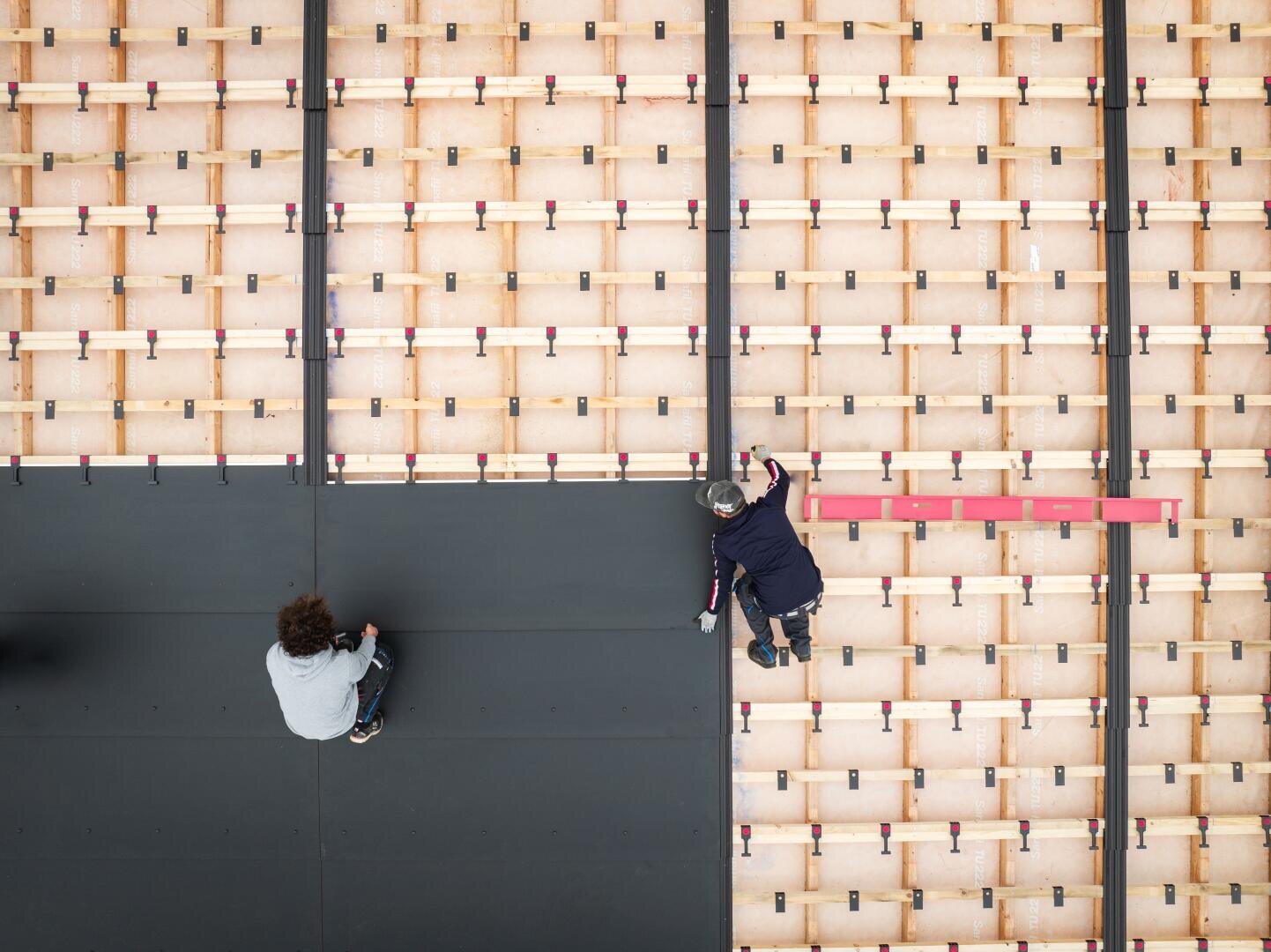 Zwei Personen arbeiten von oben gesehen an einer großen Holzkonstruktion; eine Person sitzt auf einer schwarzen Matte, die andere kniet und greift über den Rand, umgeben von einem Gitter aus Holzbrettern und roten Klammern.