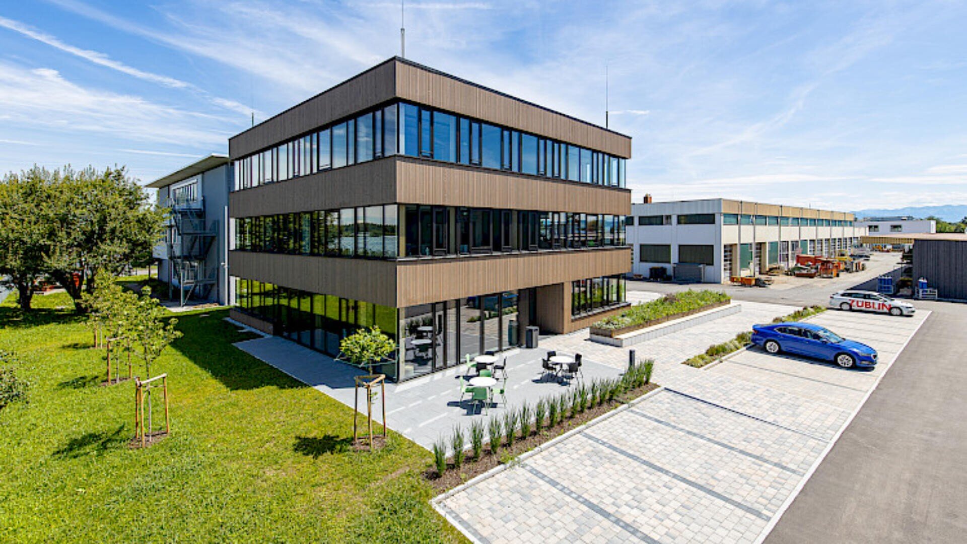 Modernes Bürogebäude mit großen Fenstern, Sitzbereich im Freien, angelegten Grünflächen und einem Parkplatz mit einigen Autos unter einem strahlend blauen Himmel mit leichter Bewölkung.