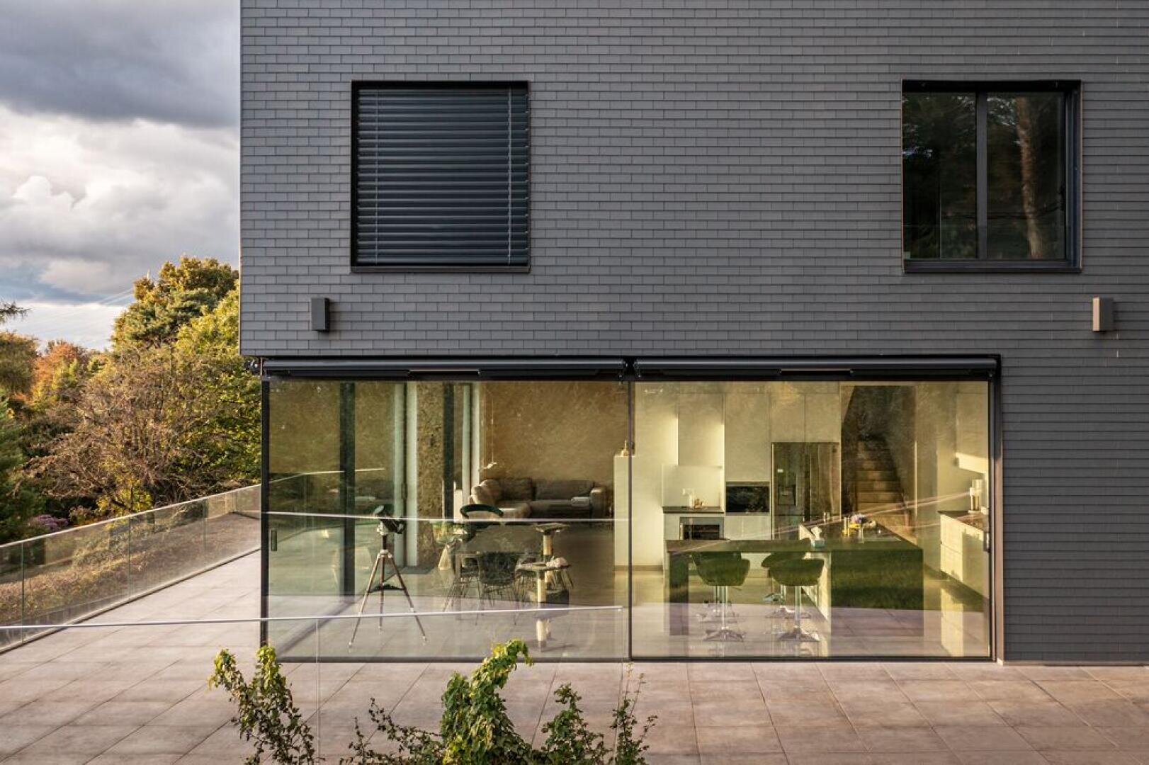 Ein modernes, graues Backsteinhaus mit großen Glasfenstern gibt den Blick frei auf eine stilvolle Küche und einen Wohnbereich im Inneren. Die geräumige Außenterrasse wird von einem Glasgeländer begrenzt, und im Hintergrund sind Bäume zu sehen.