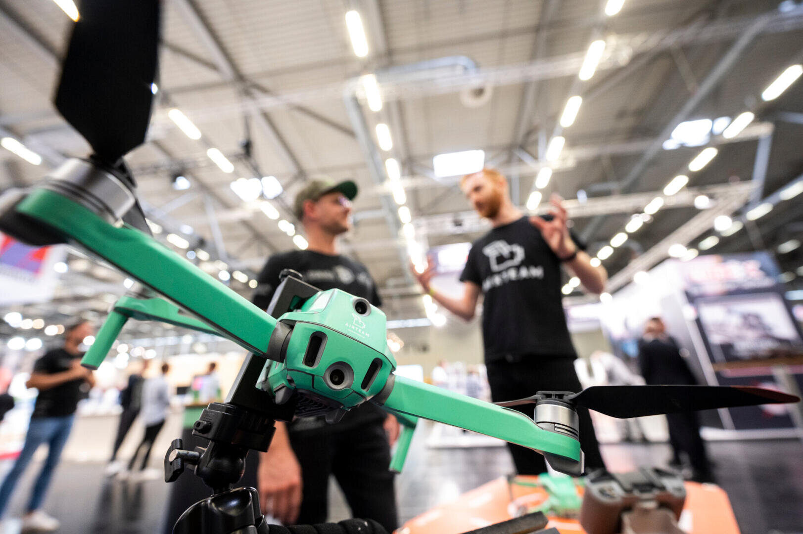 Close-up of a green drone on display indoors, with two men in black shirts and caps talking in the blurred background at an exhibition or technology event.