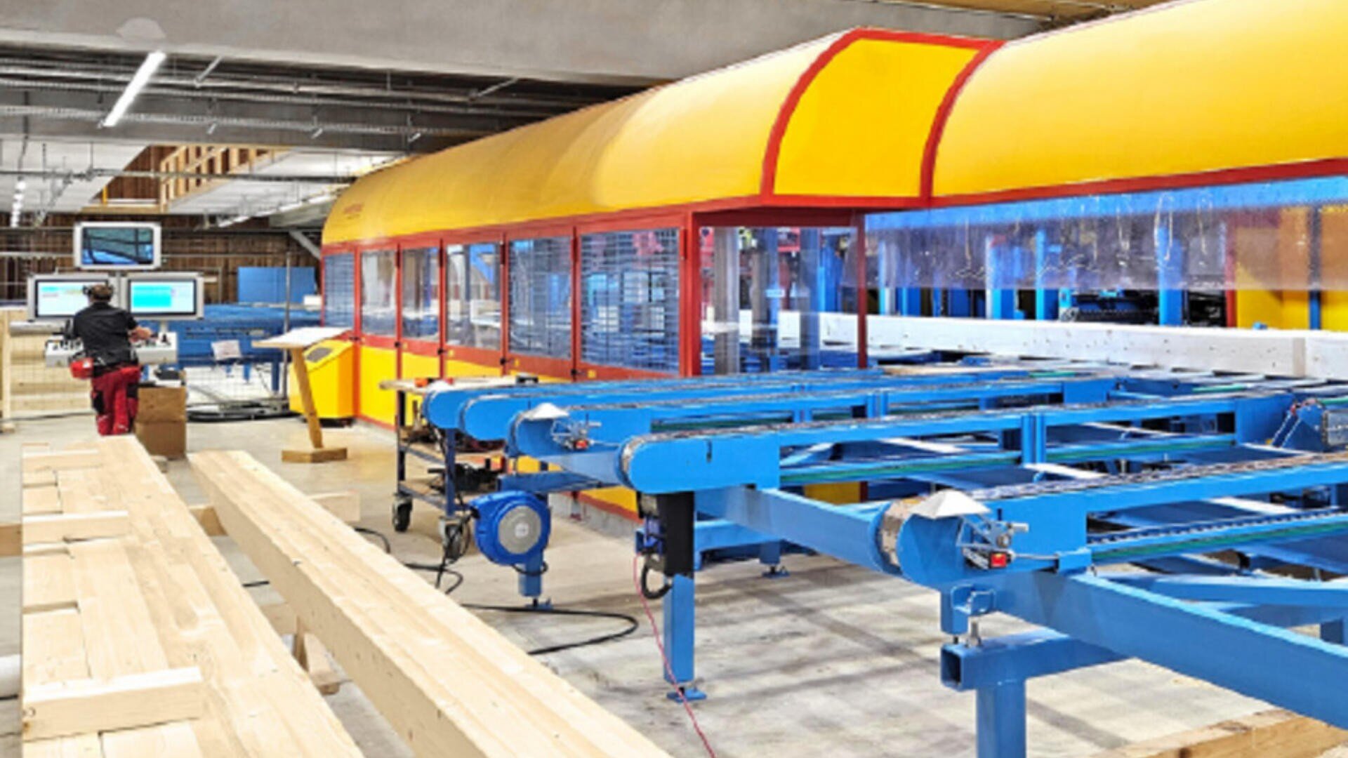 Ein Arbeiter bedient Computer neben einer großen, farbenfrohen automatisierten Maschine in einer modernen Holzverarbeitungsfabrik mit langen Holzbrettern und blauen Förderbändern.