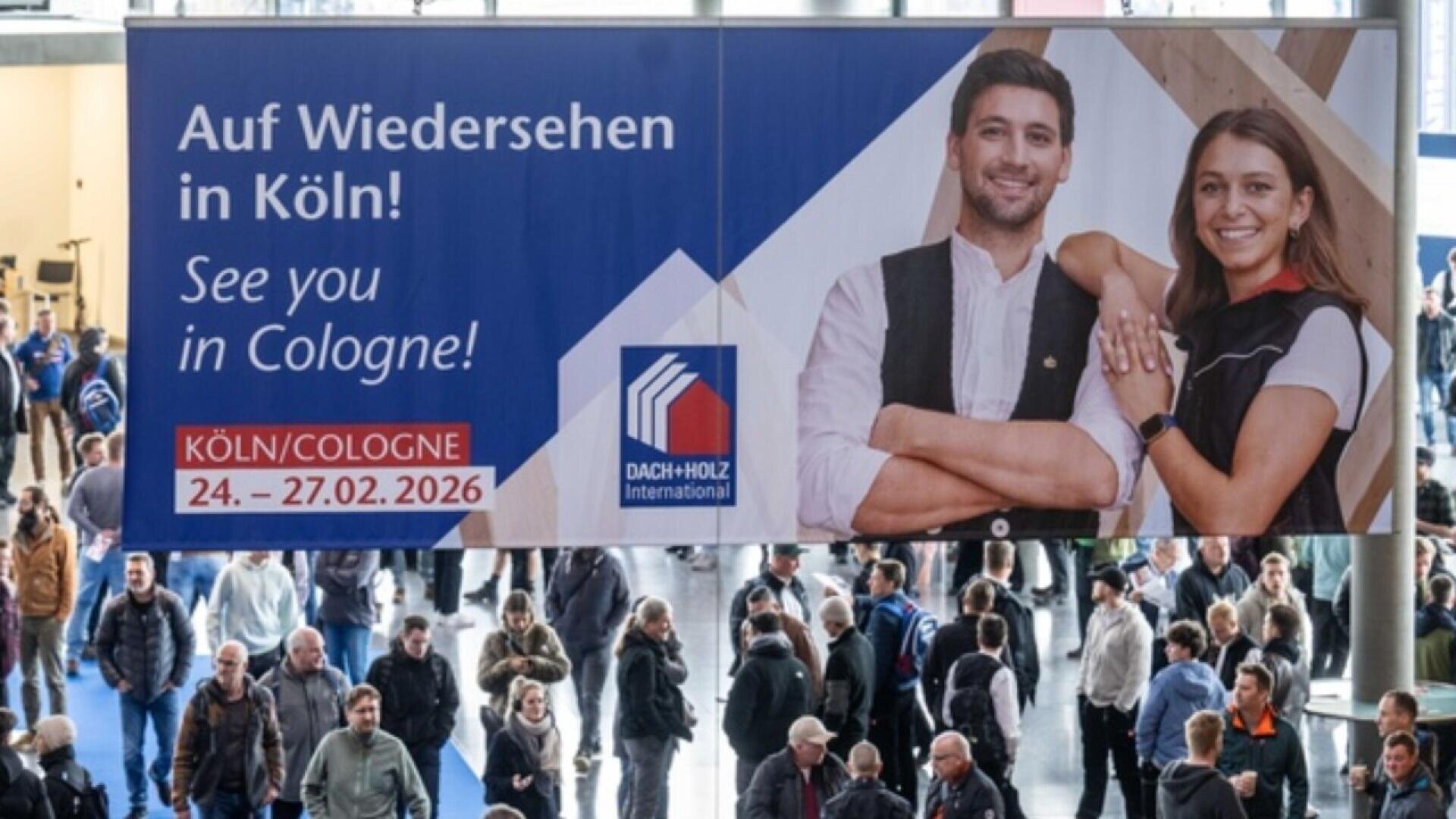 A large banner at an indoor event reads “Auf Wiedersehen in Köln! See you in Cologne!” with event dates and logos, showing a smiling man and woman. Many people gather below the banner.