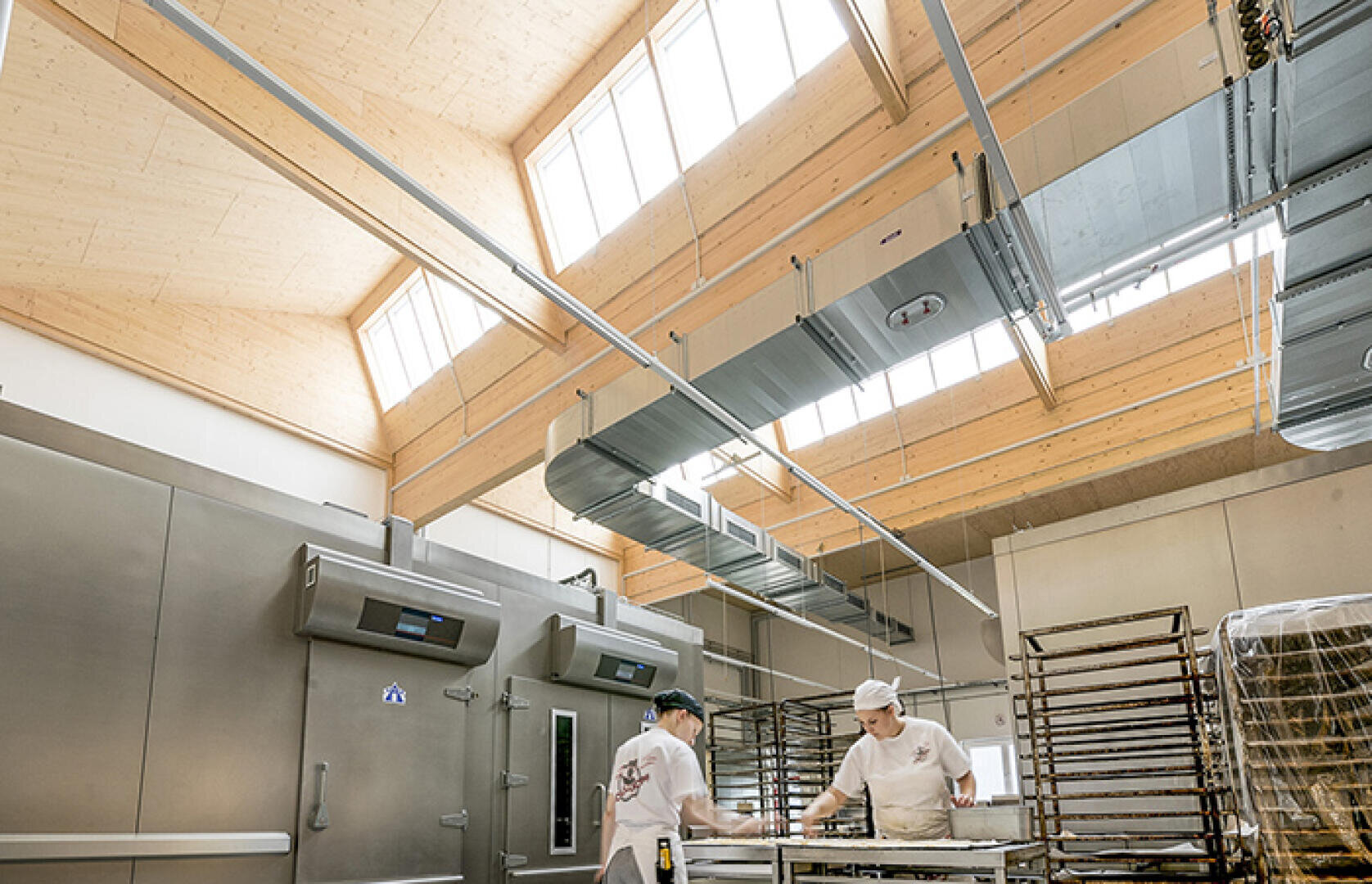 Zwei Bäcker in weißen Uniformen arbeiten an einem Tisch in einer hellen, modernen Bäckerei mit hohen Decken, großen Fenstern, Metallregalen und sichtbaren Industrieanlagen.