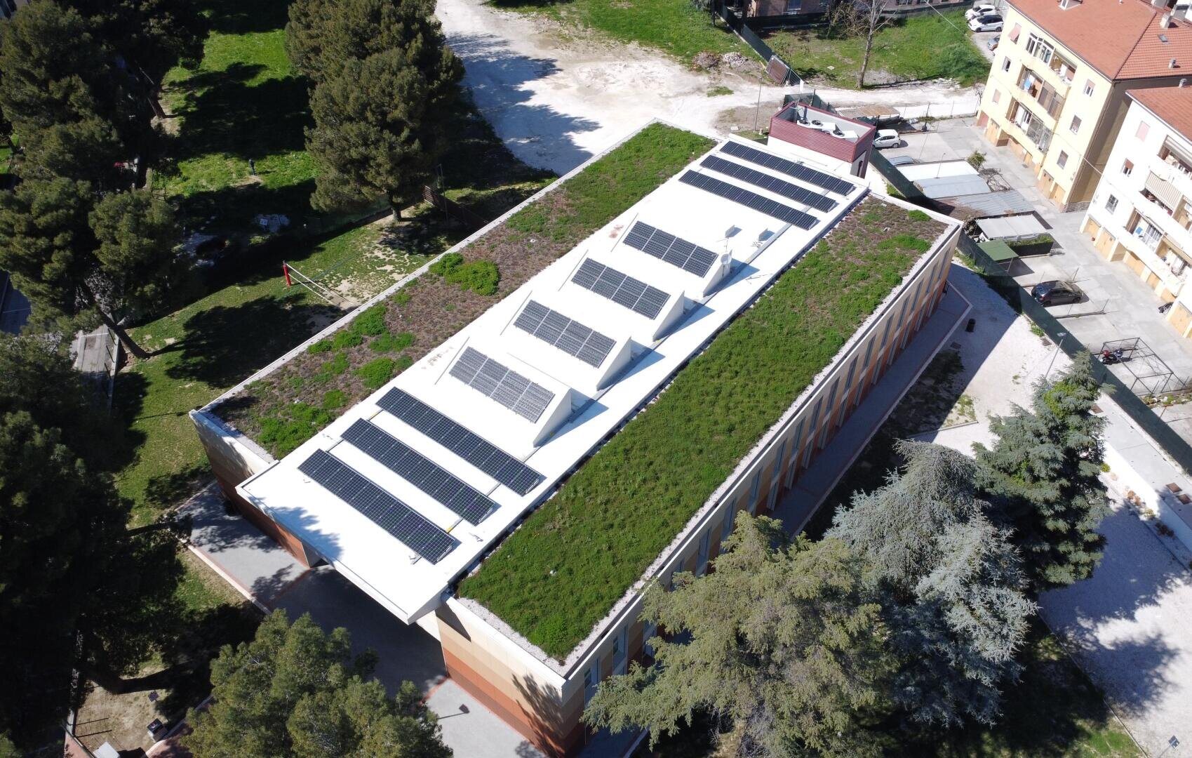 Aerial view of a rectangular building with a green roof and multiple solar panels, surrounded by trees, grassy areas, and nearby residential buildings.