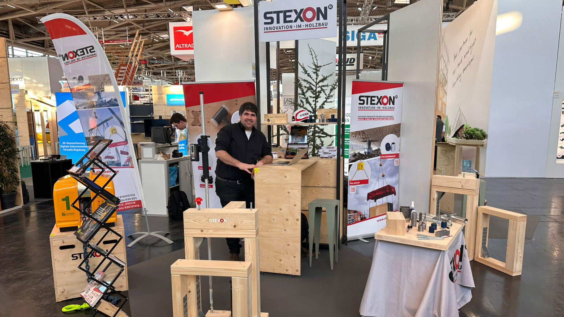 Ein Mann steht an einem hölzernen Stand, der STEXON-Werkzeuge und -Produkte auf einer Fachmesse ausstellt. Holzmöbel, Werkzeugmuster und Werbebanner sind rund um den Stand in einer großen Ausstellungshalle angeordnet.