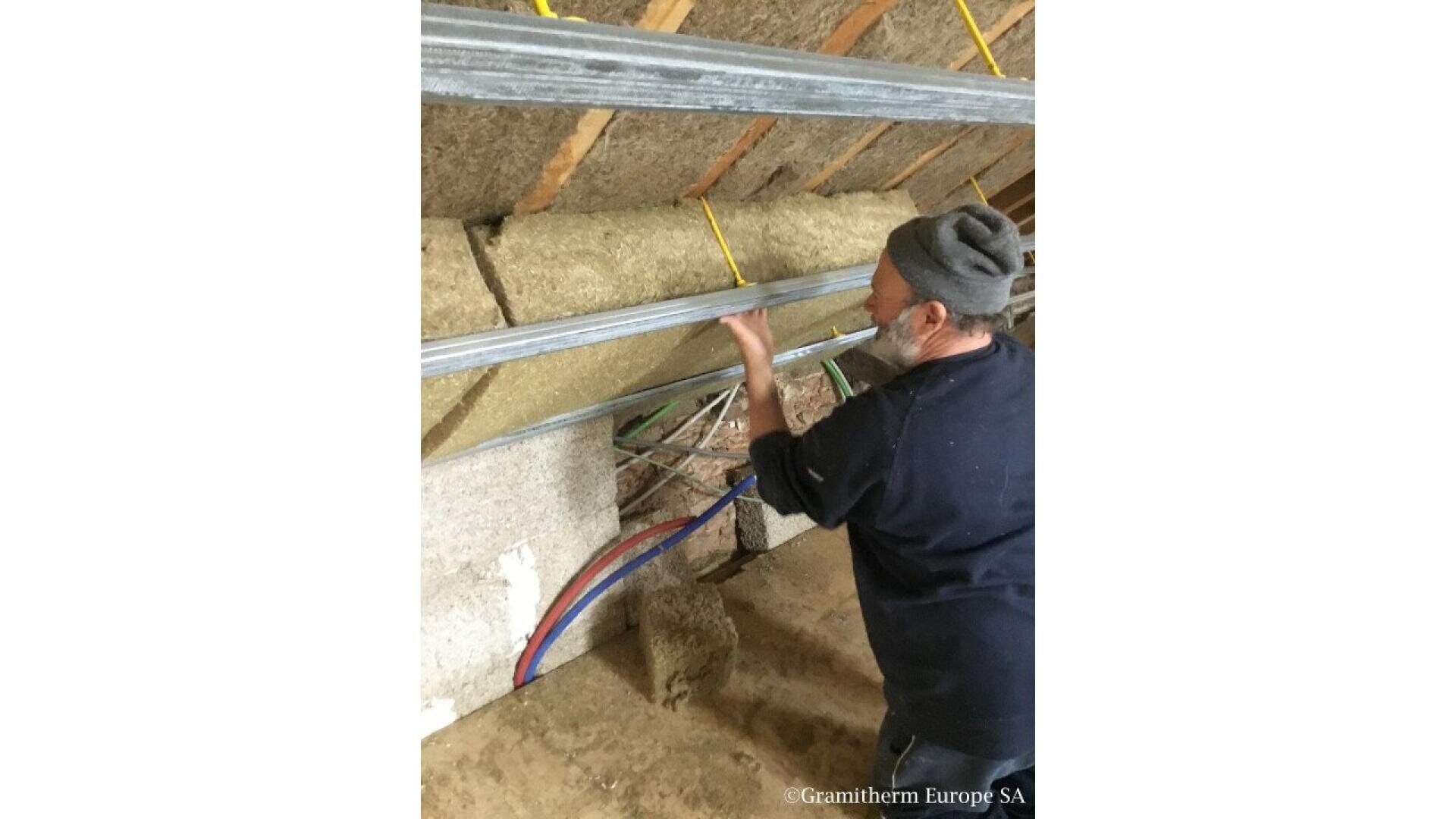 A person wearing a gray beanie installs insulation and metal framing on a wall with exposed pipes and wooden beams. The workspace appears to be under construction or renovation.