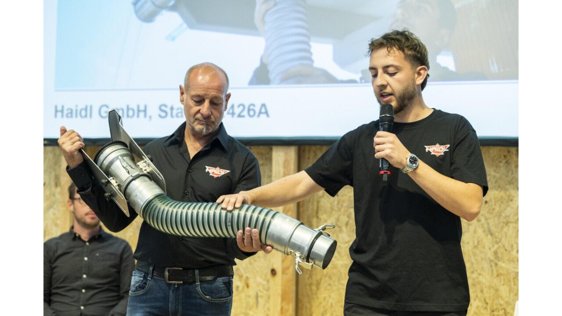Two men stand on a stage; one holds a flexible, industrial hose with metal connectors while the other speaks into a microphone and gestures toward the hose. A presentation slide is visible in the background.