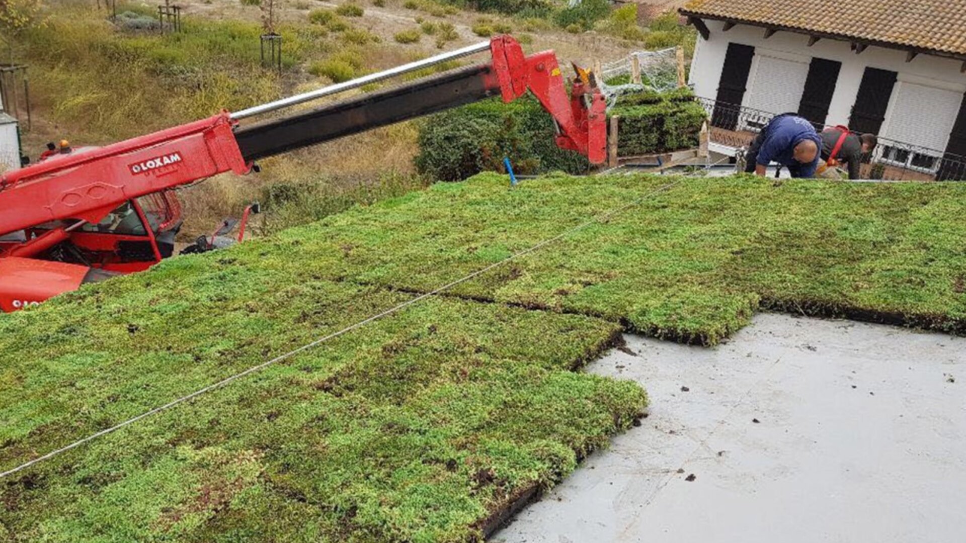 Ein Arbeiter installiert mit einem Kran Gründachplatten und legt grasbedeckte Ziegel auf ein Flachdach in der Nähe eines Hauses in einer ländlichen Gegend.