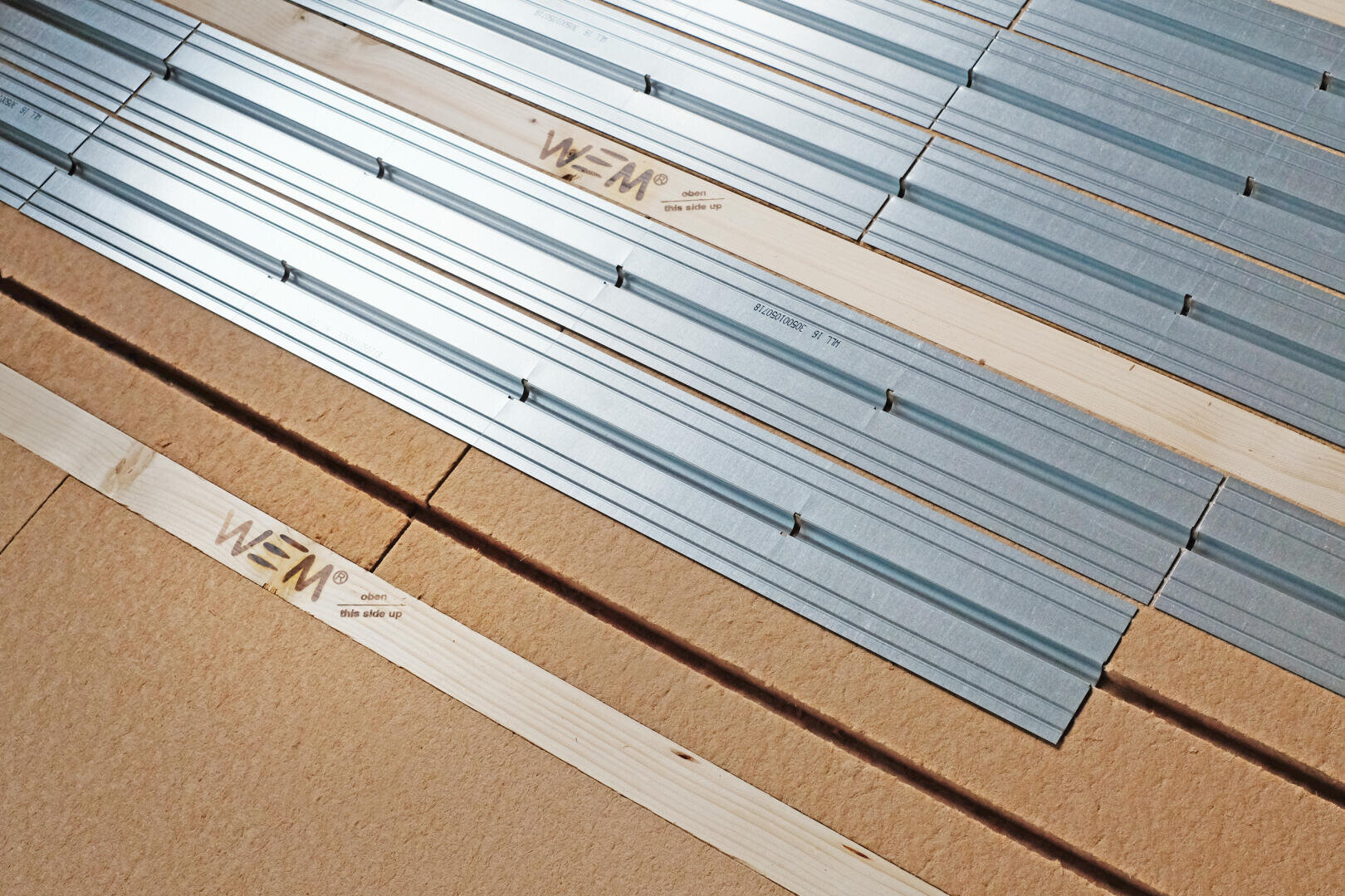 Nahaufnahme einer Bodenkonstruktion mit diagonal angeordneten Holzbrettern und Metallstreifen; das Holz ist mit dem Logo „WEM“ geprägt und die Materialien sind überlappend verlegt.