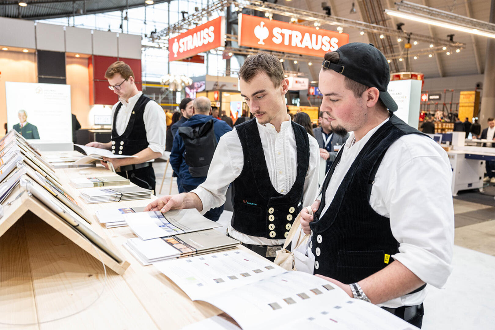 Drei junge Männer in traditionellen Arbeitswesten prüfen Broschüren an einem Ausstellungstisch in einer belebten Messehalle, im Hintergrund sind Strauss-Schilder zu sehen.