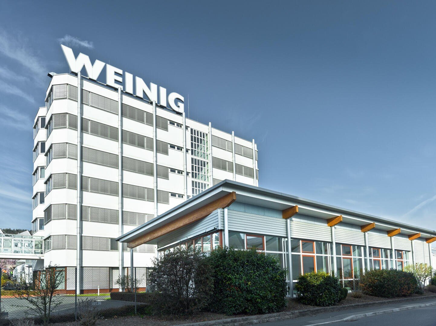 Ein modernes, mehrstöckiges Gebäude mit großen WEINIG-Schriftzügen auf dem Dach steht hinter einer niedrigen, von Büschen gesäumten Glasfront unter einem strahlend blauen Himmel.