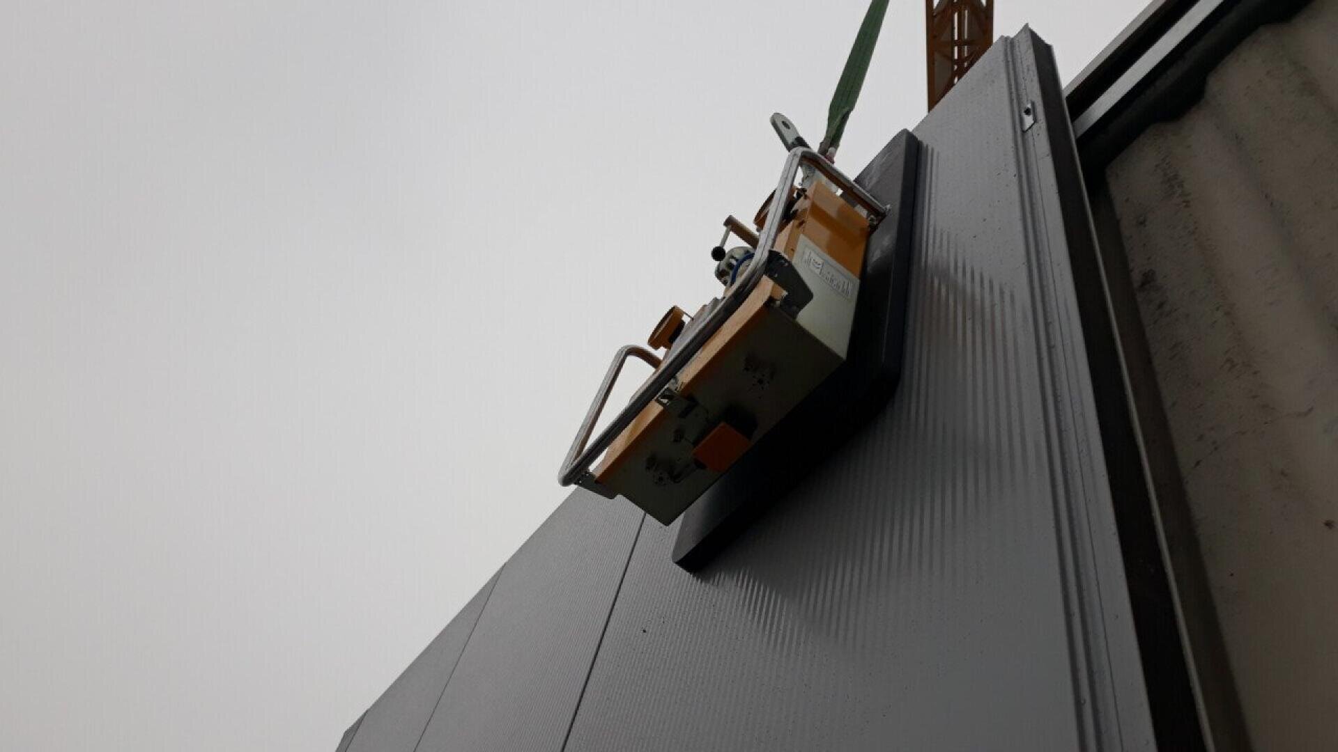 Eine Nahaufnahme eines Krans, der eine große graue Metallplatte mit einem Saugnapfheber anhebt, von unten vor einem bewölkten Himmel gesehen.