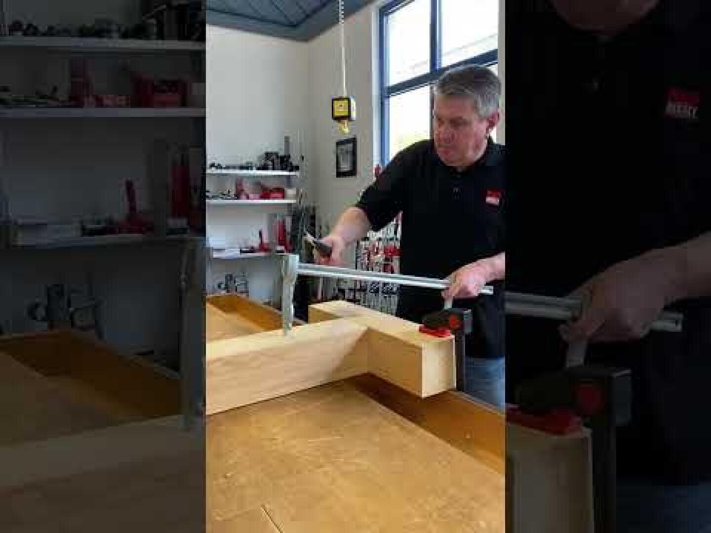 Eine Person in einer Werkstatt benutzt eine große Zwinge, um zwei Holzstücke auf einer Werkbank zu befestigen, wobei im Hintergrund Werkzeuge und Regale zu sehen sind.