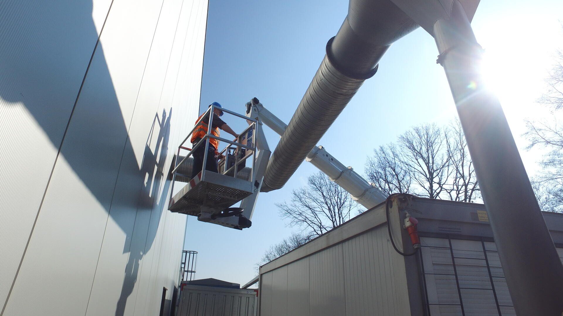 Zwei Arbeiter in Schutzkleidung stehen auf einer Hebebühne neben einem großen Industriegebäude und arbeiten an einem sonnigen Tag in der Nähe eines großen Lüftungsrohrs, während im Hintergrund blattlose Bäume zu sehen sind.