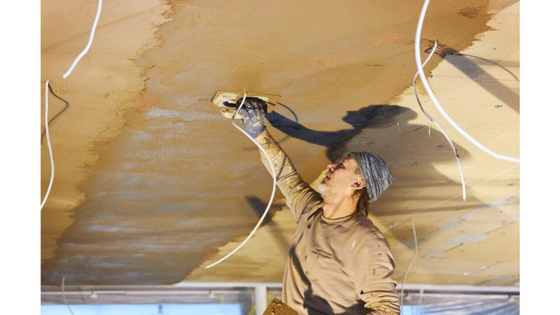 A construction worker wearing a gray headscarf and gloves smooths plaster on a ceiling with a trowel. Loose white wires hang down around him, and sunlight illuminates the workspace.
