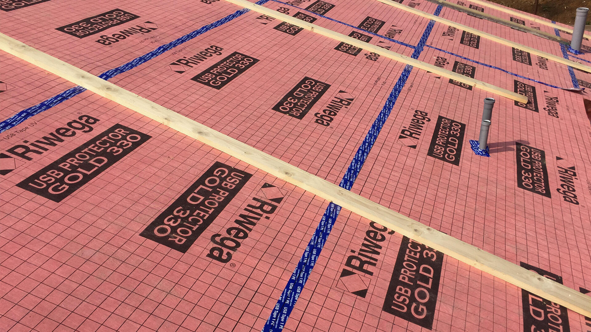 Close-up of a roof under construction covered with a pink waterproof membrane labeled Riwega USB Protector Gold 330, with wooden battens and several pipes protruding through the surface.