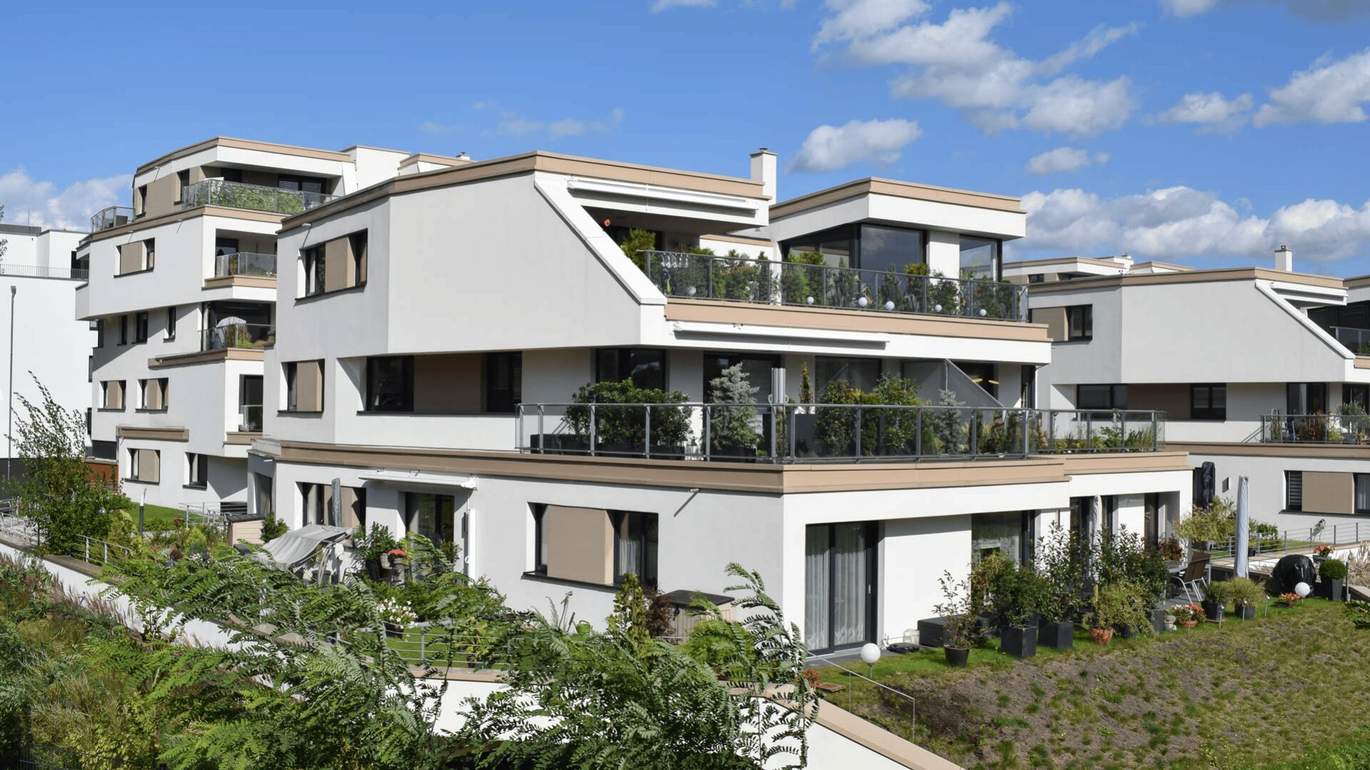 Moderne weiße Wohnhäuser mit großen Balkonen und Glasgeländern, umgeben von Grün und Pflanzen, unter einem blauen Himmel mit vereinzelten Wolken.