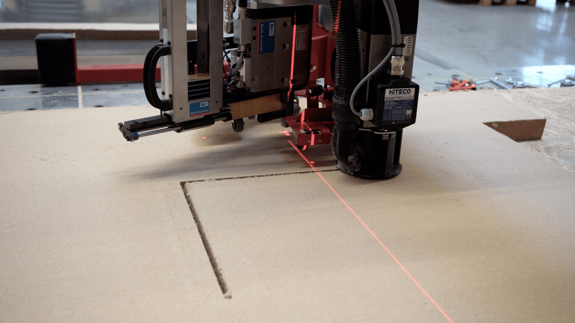 In einer industriellen Werkstatt schneidet eine CNC-Maschine mit einem sichtbaren roten Laser eine rechteckige Nut in eine große Materialplatte, wahrscheinlich Holz oder MDF.