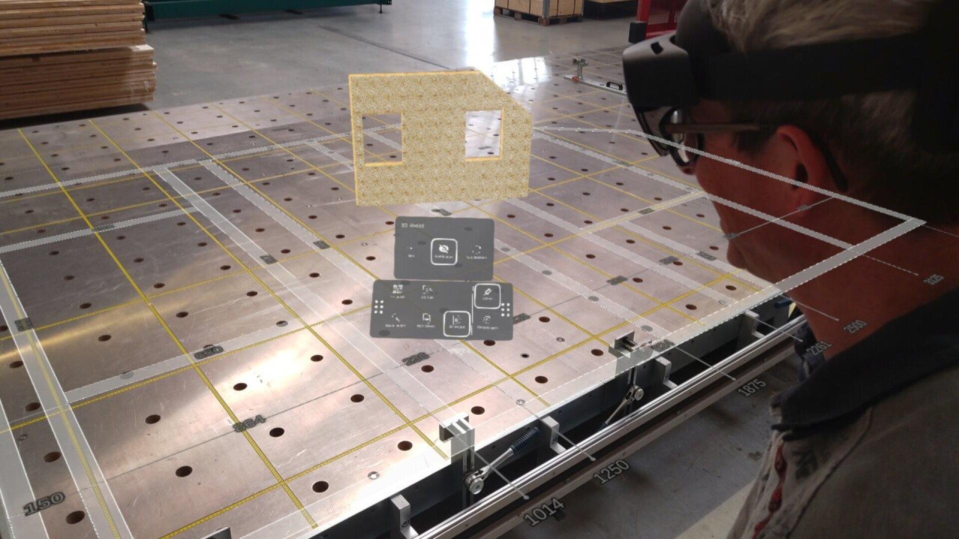 A person wearing augmented reality glasses views a virtual interface and 3D structure projected over a large metal workbench with a grid and measurement markings in an industrial setting.